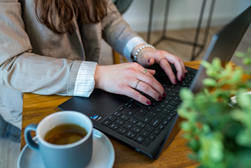 Hände einer Person bei der vorbereitenden Buchhaltung am Laptop, mit einer Tasse Kaffee auf einem Holztisch in ruhiger Arbeitsumgebung.